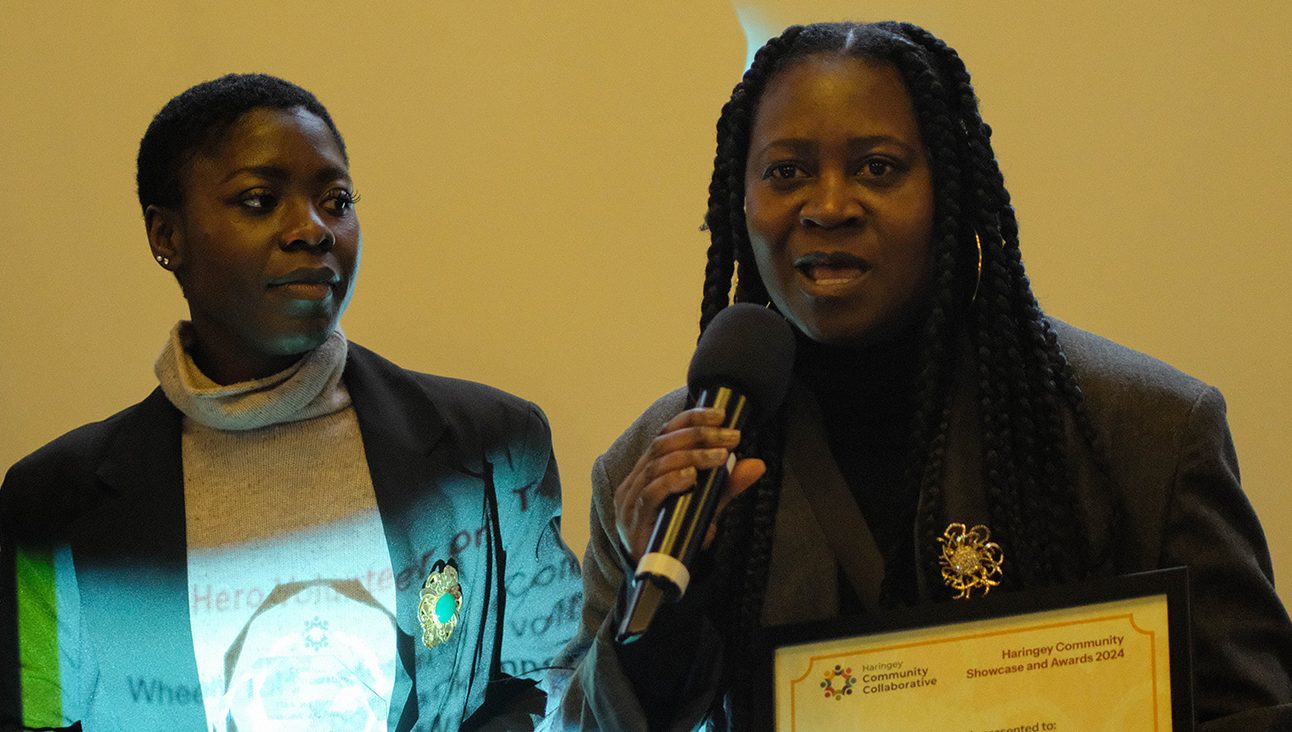 Two black woman on a stage. One is speaking into a microphone while holding an award