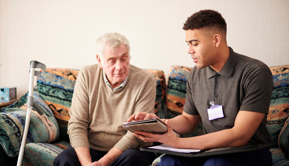 Mixed heritage support worker providing one to one support and advice to older white man in his home, with both people sitting on a sofa.