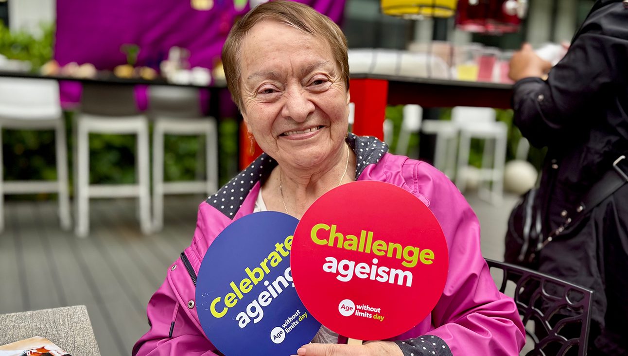 An older woman smiling whilst holding up two signs. One reads 'celebrate ageing', and the other reads 'challenge ageism'.