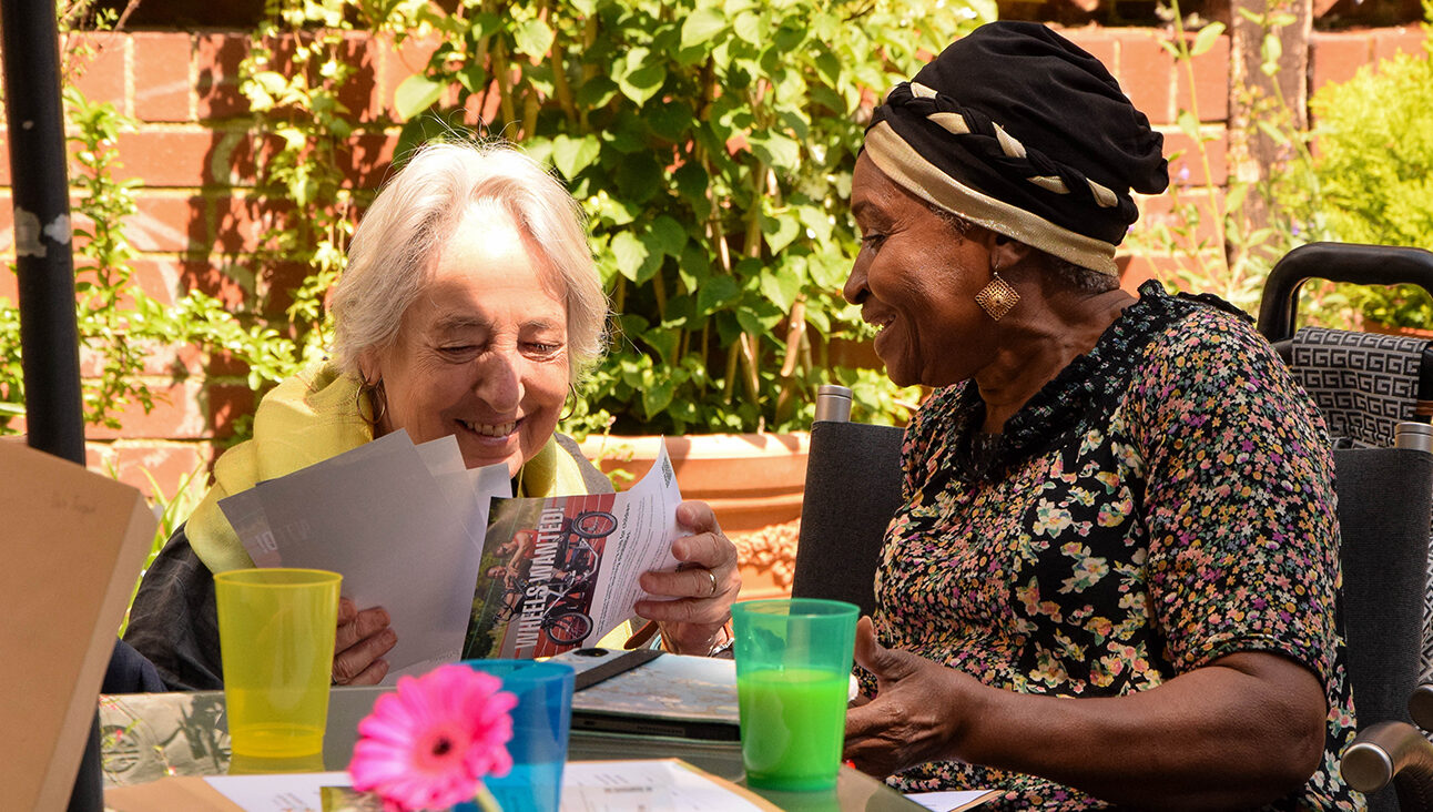 An old white woman and an old black woman having a conversation at at table where there are cups of juice. It is a sunny day and they are both under a parasol.