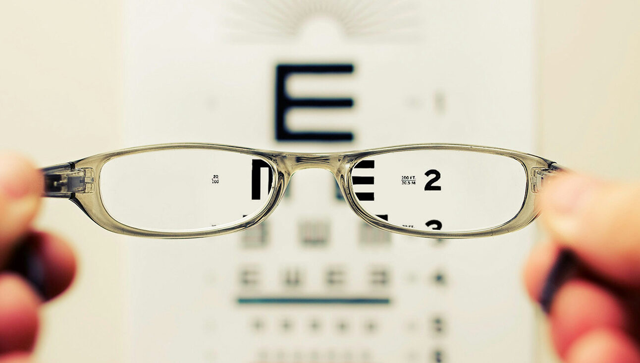 A pair of hands holding up a paid of glasses in front of an eye test chart. The lenses of the glasses make the eye chart in focus