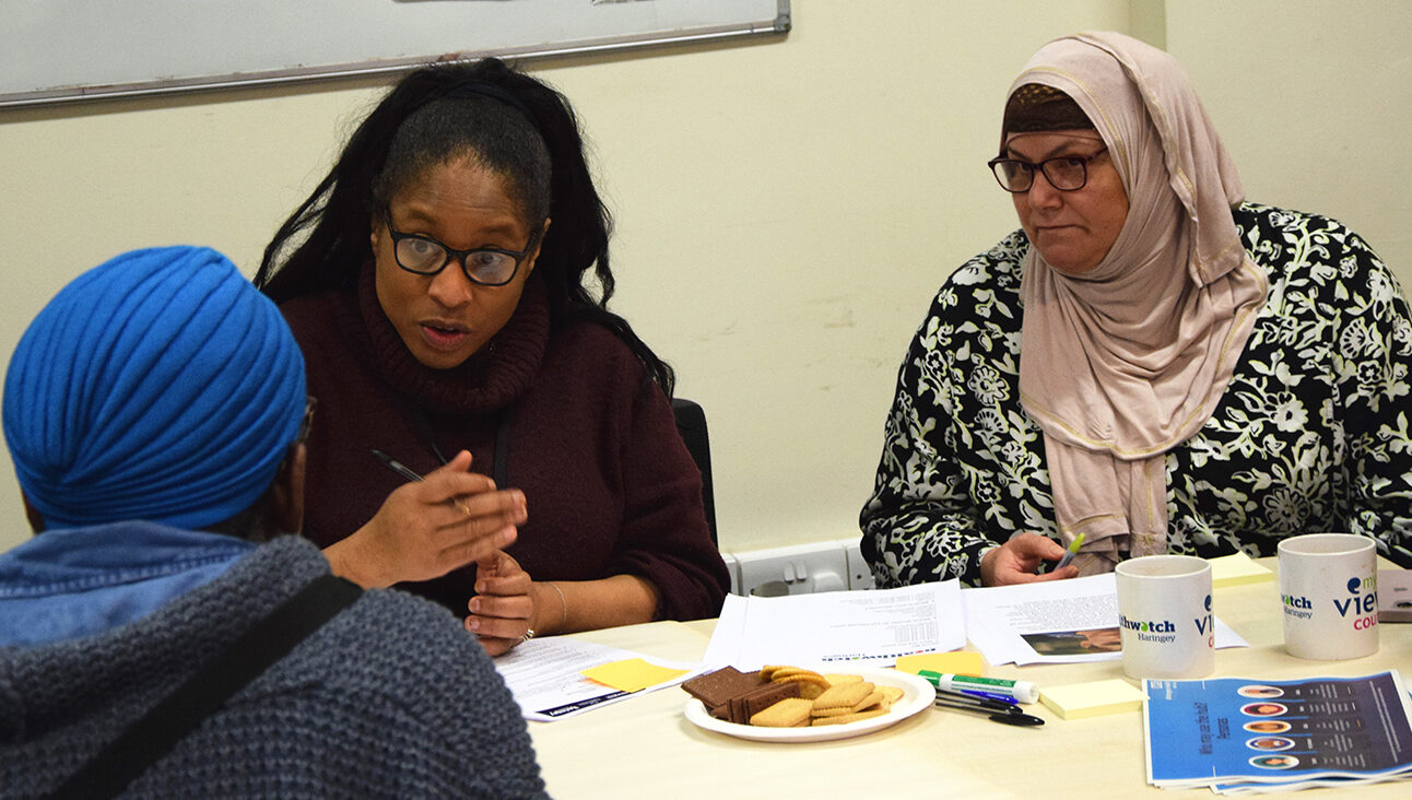 Two black women, one who is wearing a blue hat, and a Muslim women with her head covered have a conversation at a table. There are papers, pens and biscuits on the table.